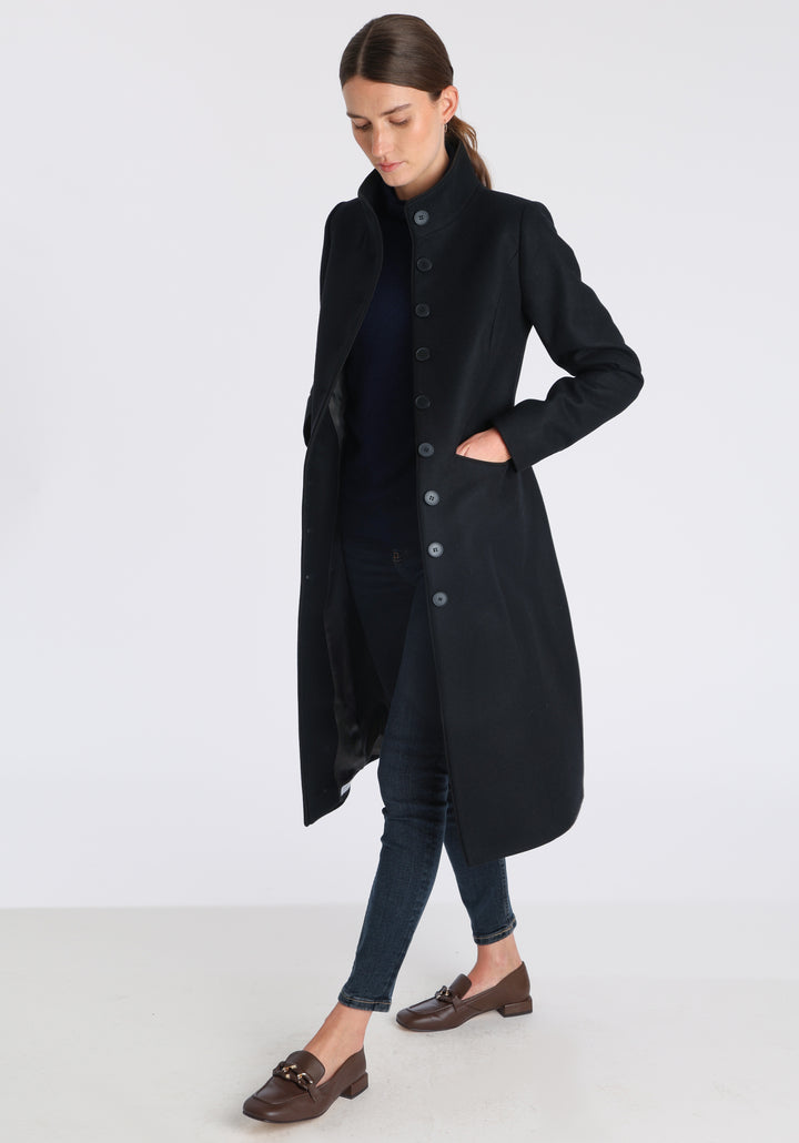 Model wearing the Opera coat in black cashmere blend, an elegant, knee-length style with a high funnel neck, curved front pockets, and a slim, waist-contouring silhouette.