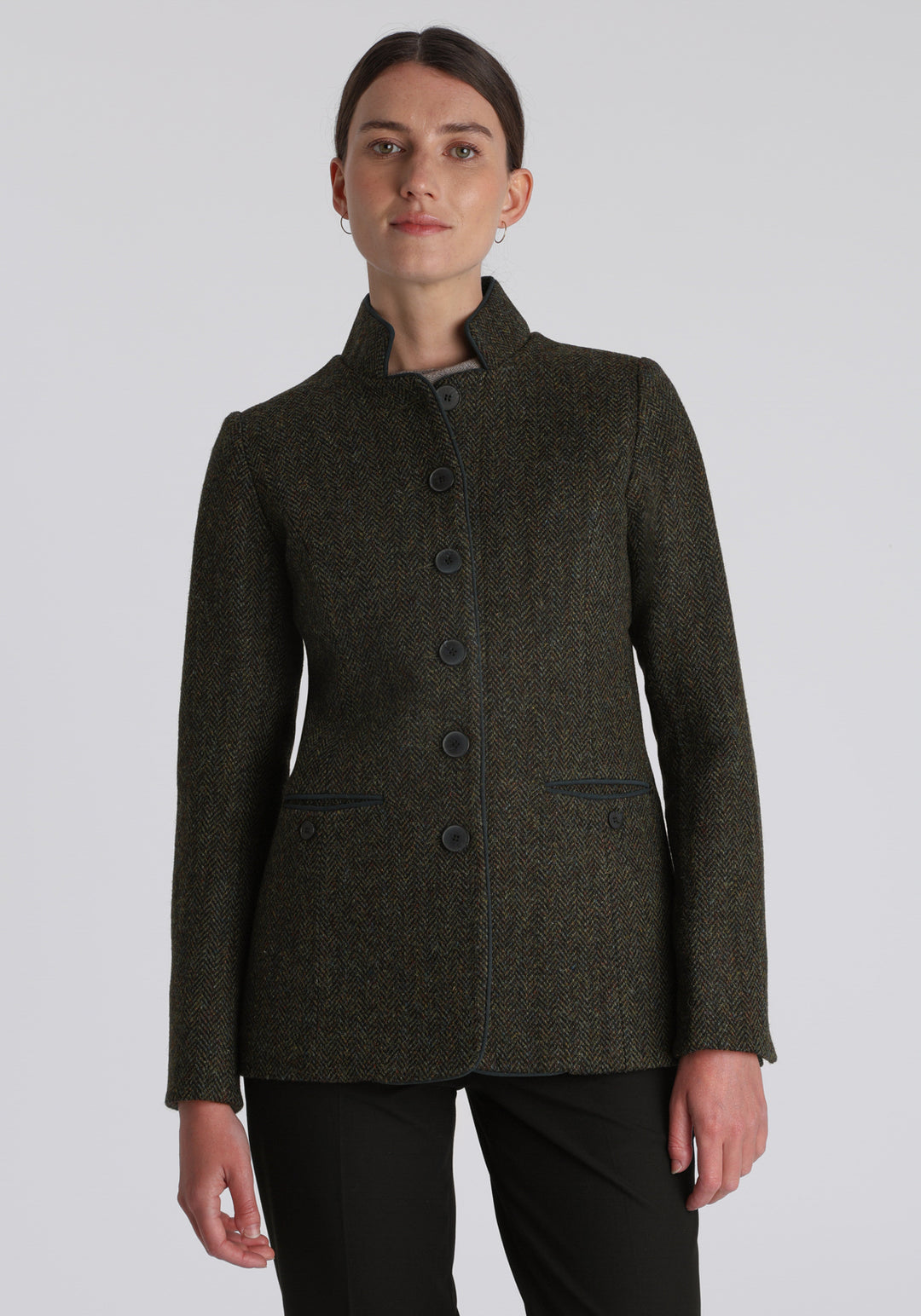 Model wearing the Patmos jacket in green spruce herringbone Harris Tweed with suede trim, a slim-fit style featuring a mandarin collar, front pockets, and generous sleeves with slate button detailing.