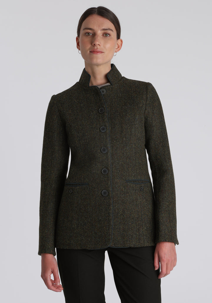 Model wearing the Patmos jacket in green spruce herringbone Harris Tweed with suede trim, a slim-fit style featuring a mandarin collar, front pockets, and generous sleeves with slate button detailing.