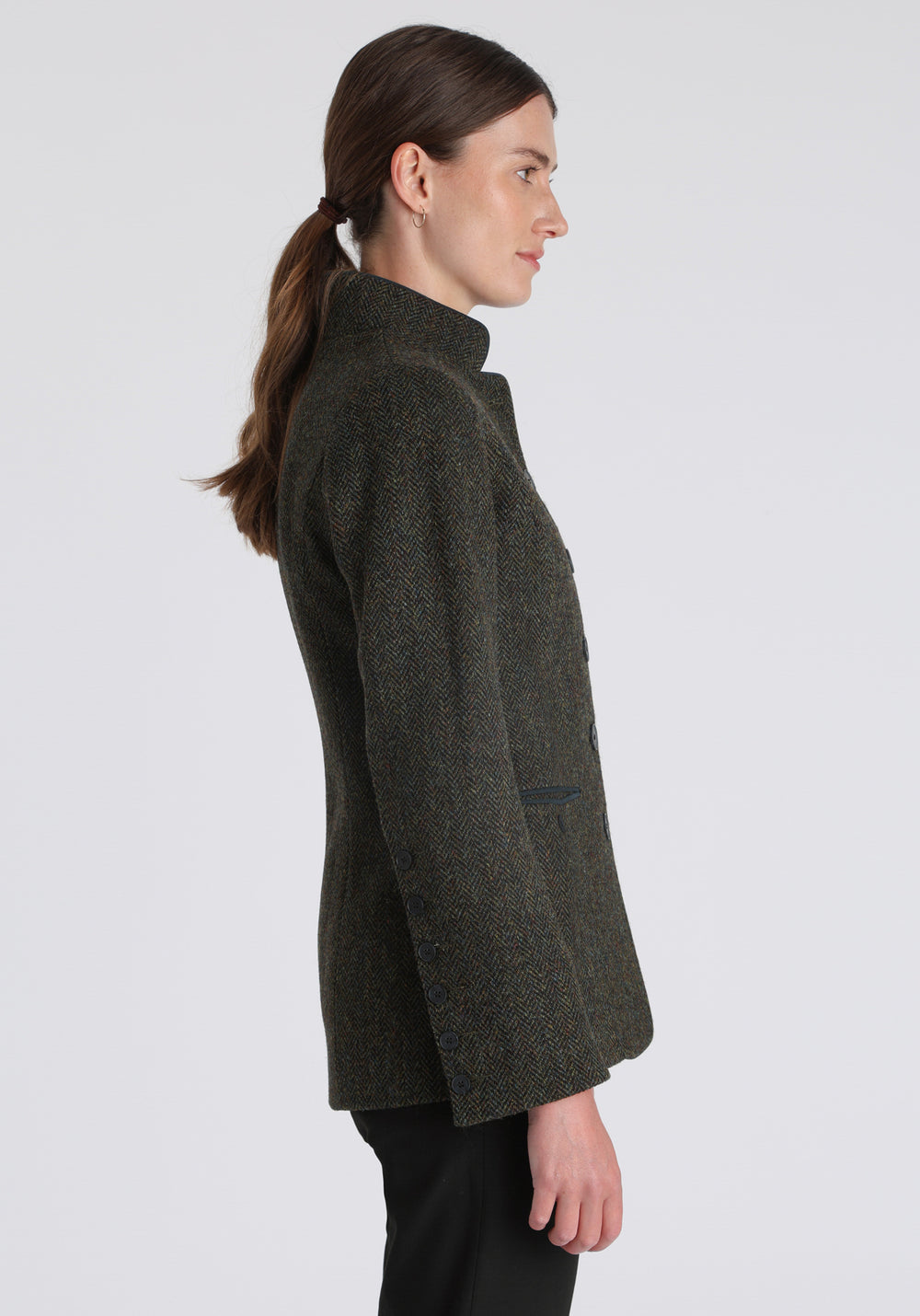 Model wearing the Patmos jacket in green spruce herringbone Harris Tweed with suede trim, a slim-fit style featuring a mandarin collar, front pockets, and generous sleeves with slate button detailing.