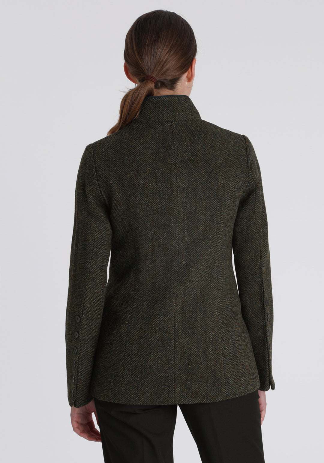 Model wearing the Patmos jacket in green spruce herringbone Harris Tweed with suede trim, a slim-fit style featuring a mandarin collar, front pockets, and generous sleeves with slate button detailing.