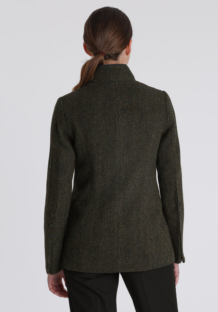 Model wearing the Patmos jacket in green spruce herringbone Harris Tweed with suede trim, a slim-fit style featuring a mandarin collar, front pockets, and generous sleeves with slate button detailing.