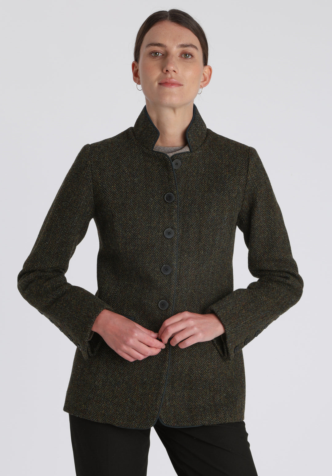 Model wearing the Patmos jacket in green spruce herringbone Harris Tweed with suede trim, a slim-fit style featuring a mandarin collar, front pockets, and generous sleeves with slate button detailing.
