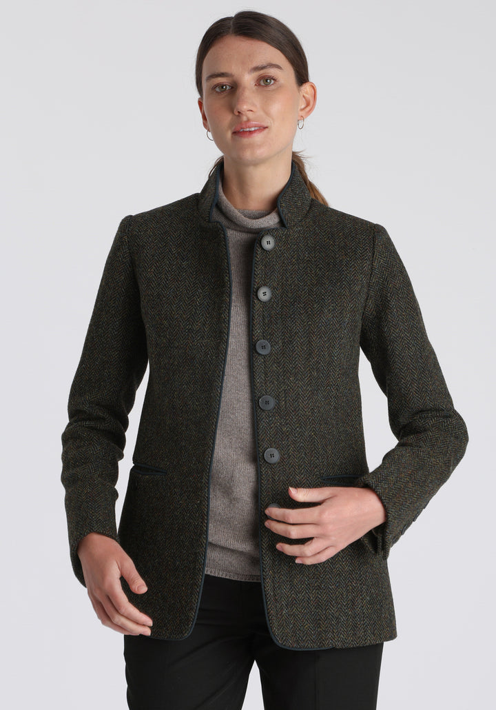 Model wearing the Patmos jacket in green spruce herringbone Harris Tweed with suede trim, a slim-fit style featuring a mandarin collar, front pockets, and generous sleeves with slate button detailing.