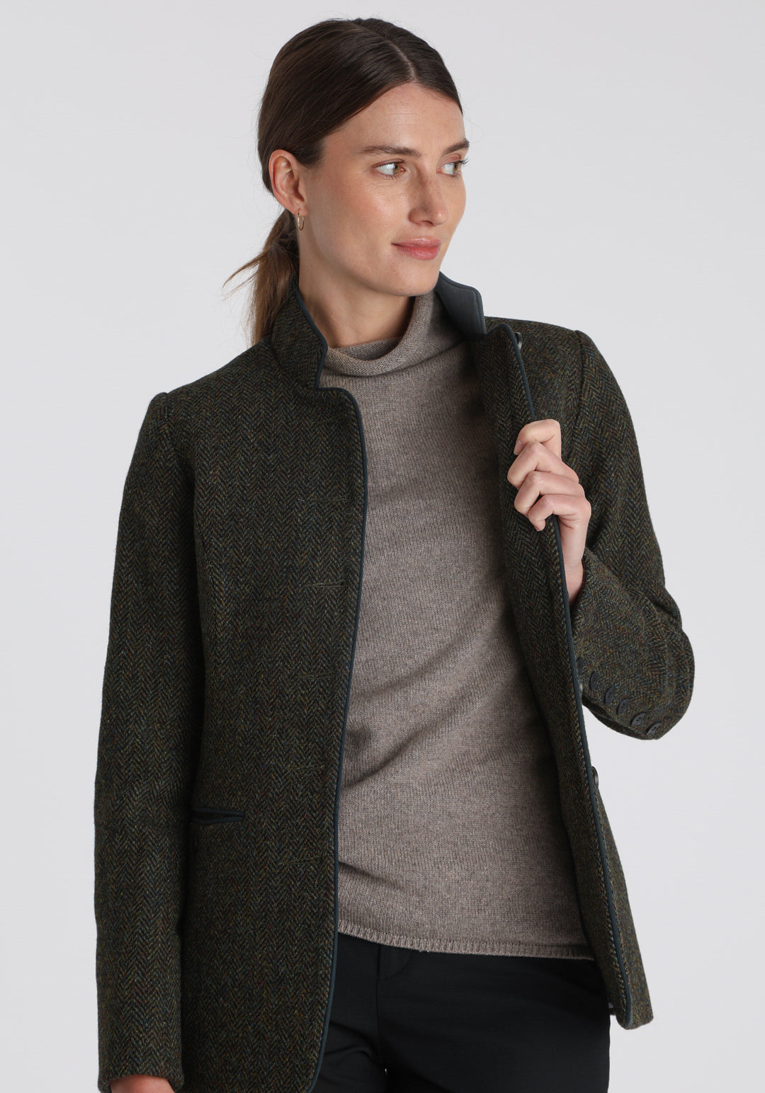 Model wearing the Patmos jacket in green spruce herringbone Harris Tweed with suede trim, a slim-fit style featuring a mandarin collar, front pockets, and generous sleeves with slate button detailing.