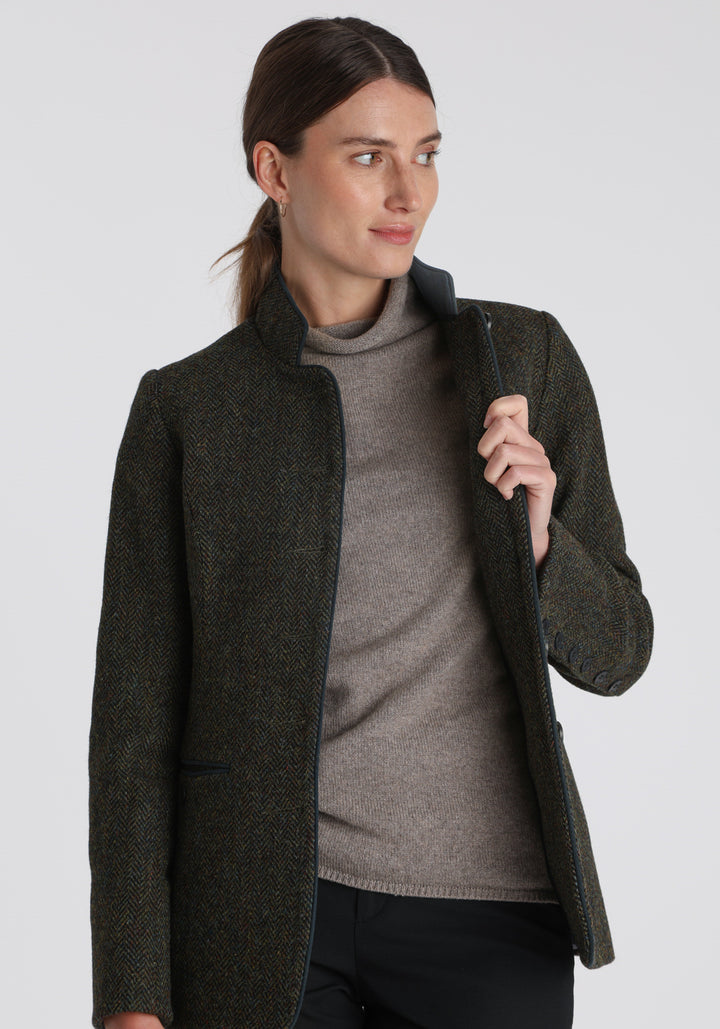 Model wearing the Patmos jacket in green spruce herringbone Harris Tweed with suede trim, a slim-fit style featuring a mandarin collar, front pockets, and generous sleeves with slate button detailing.