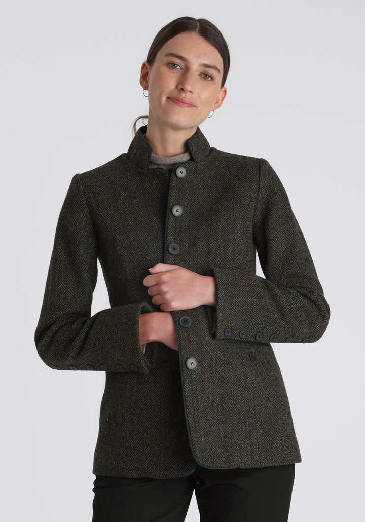 Model wearing the Patmos jacket in green spruce herringbone Harris Tweed with suede trim, a slim-fit style featuring a mandarin collar, front pockets, and generous sleeves with slate button detailing.