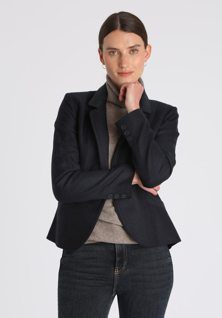 Model wearing the Tallulah jacket in navy herringbone lambswool with suede trim, a tailored style with a single button fastening and a collar that can be worn turned up or down.