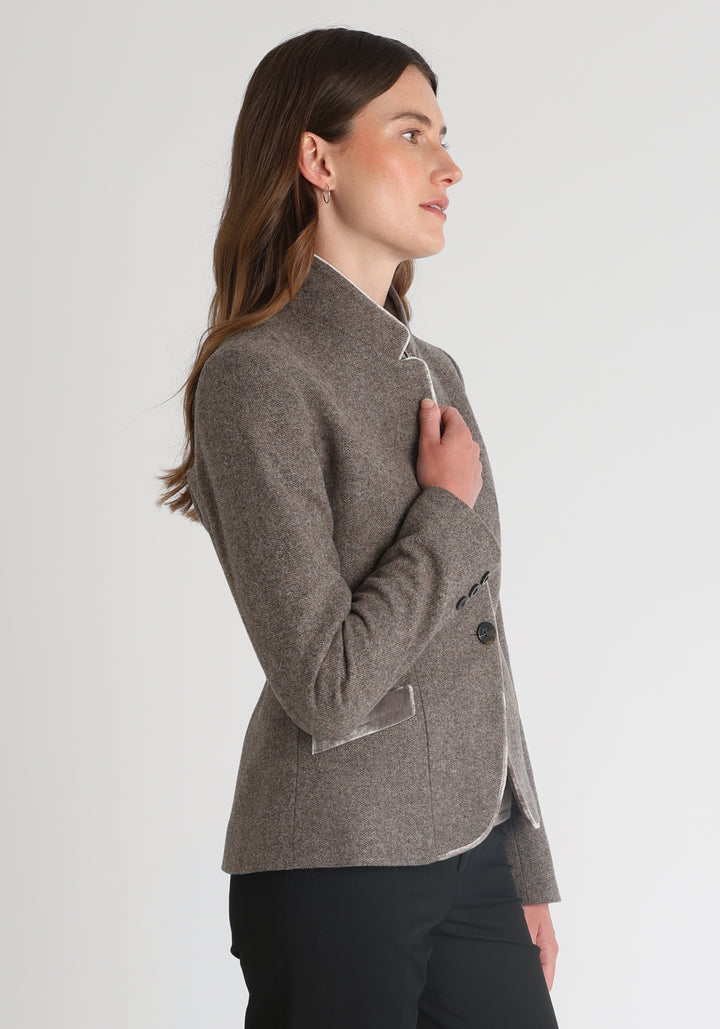 Model wearing the Tallulah jacket in a luxuriously soft cool mallow taupe herringbone lambswool with silk velvet trim, a tailored style with a single button fastening and a collar that can be worn turned up or down.
