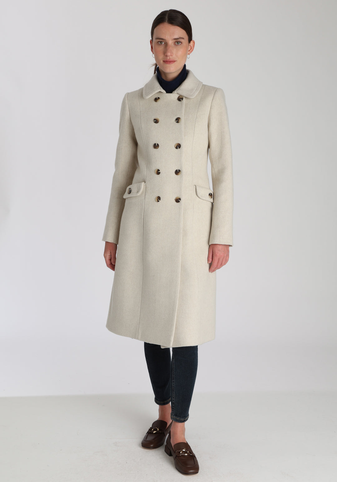 Model wearing the Tori coat in cream merino wool, a vintage-inspired style with a double-breasted front, gently flared skirt, and curved detailing on the collar, pockets, and back belt.