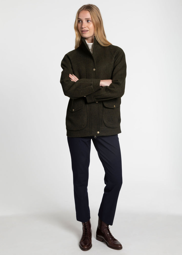 A woman modeling a dark olive green wool jacket with front flap pockets, paired with navy trousers and brown leather boots, against a plain light gray studio background.