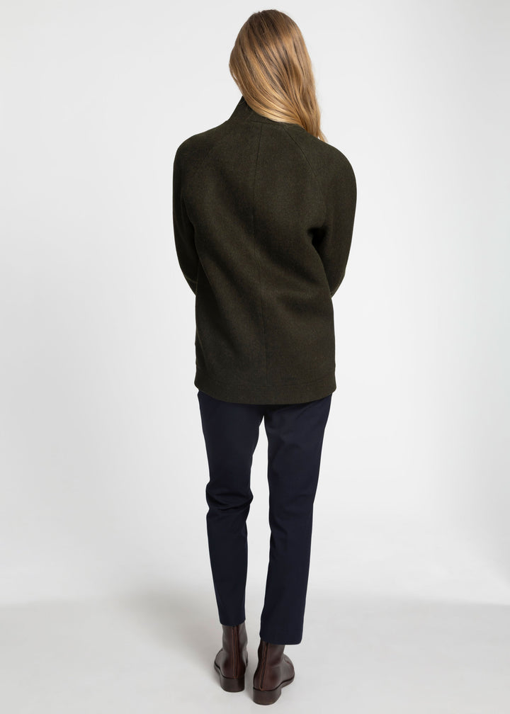 A woman viewed from behind wearing a dark olive green wool jacket, navy blue pants, and brown leather boots against a plain white studio background.