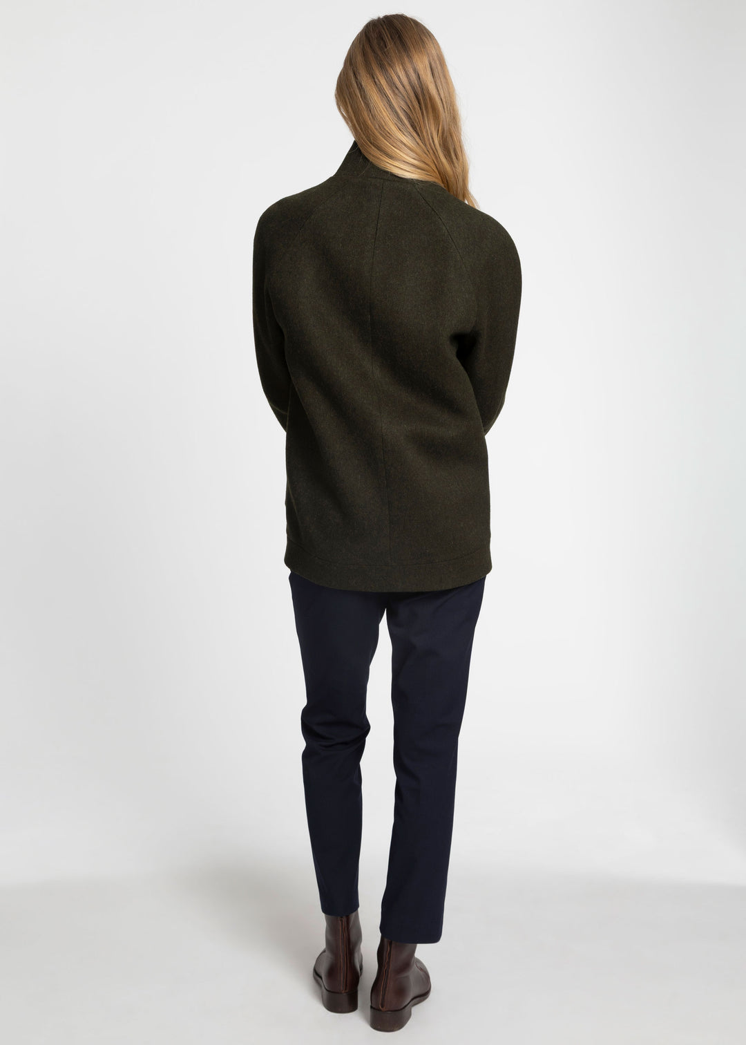 A woman viewed from behind wearing a dark olive green wool jacket, navy blue pants, and brown leather boots against a plain white studio background.