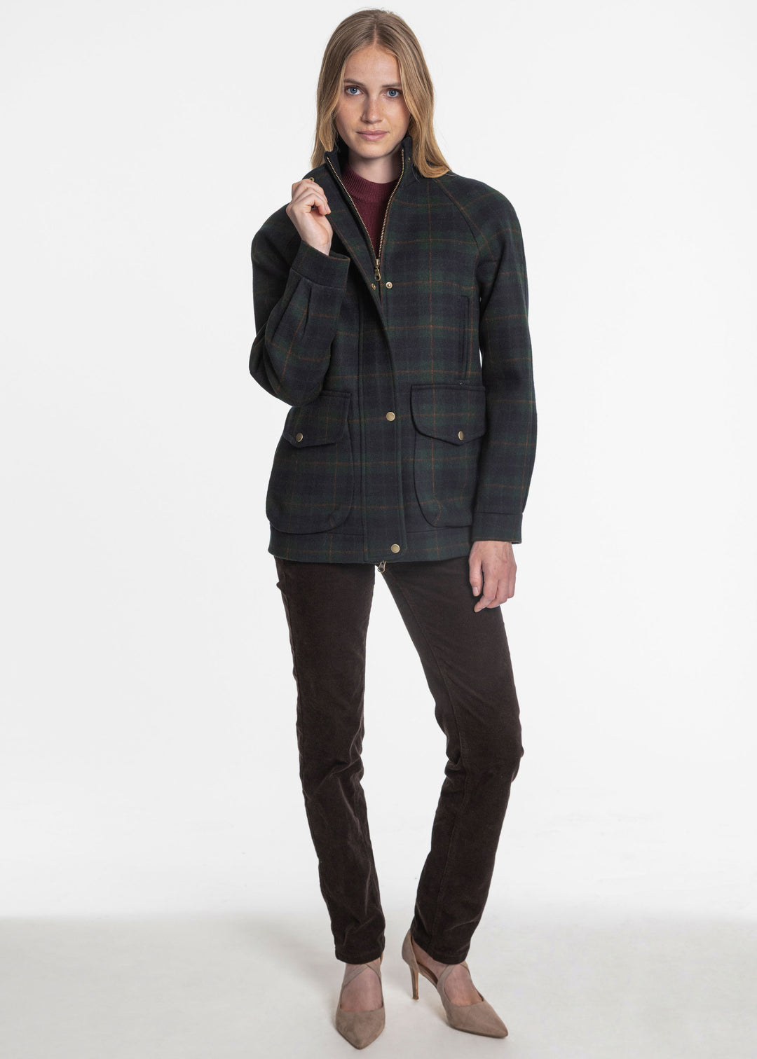 A woman modeling a dark green and navy plaid jacket with bronze snap buttons, paired with dark brown pants and beige heels, against a plain white studio background.