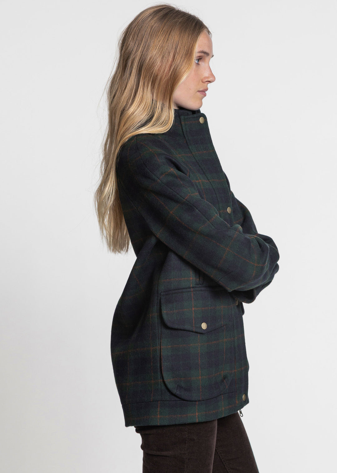 A woman shown in profile wearing a dark green and navy plaid jacket with bronze buttons, set against a plain light gray studio background.