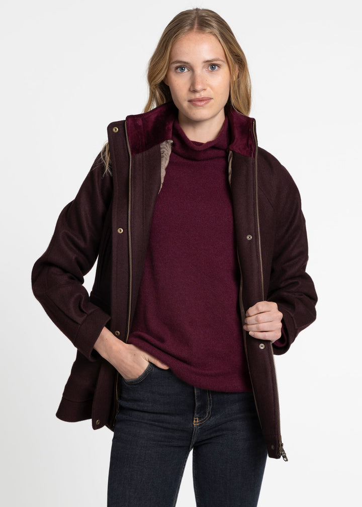 A woman modeling a dark burgundy jacket with a plush velvet-like collar lining over a matching burgundy turtleneck sweater and dark blue jeans, shot against a plain white studio background.