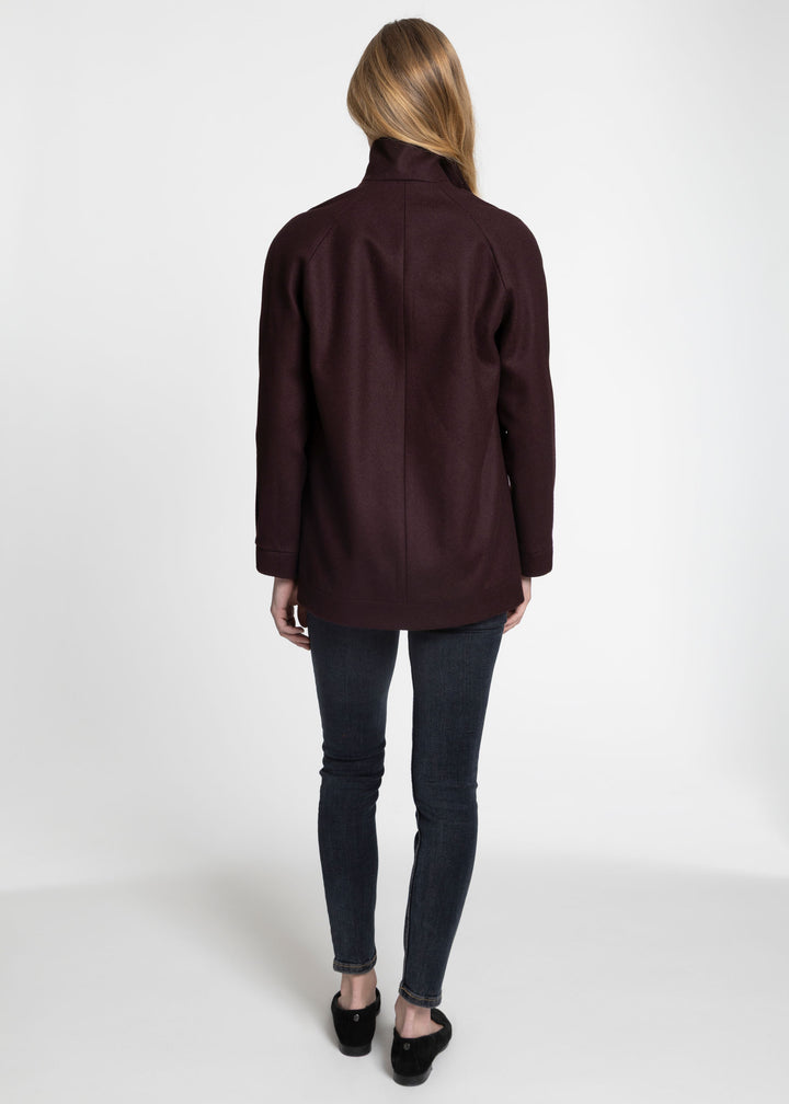 A woman viewed from the back wearing a dark burgundy long-sleeve jacket, dark gray skinny jeans, and black flats, standing against a plain white studio background.