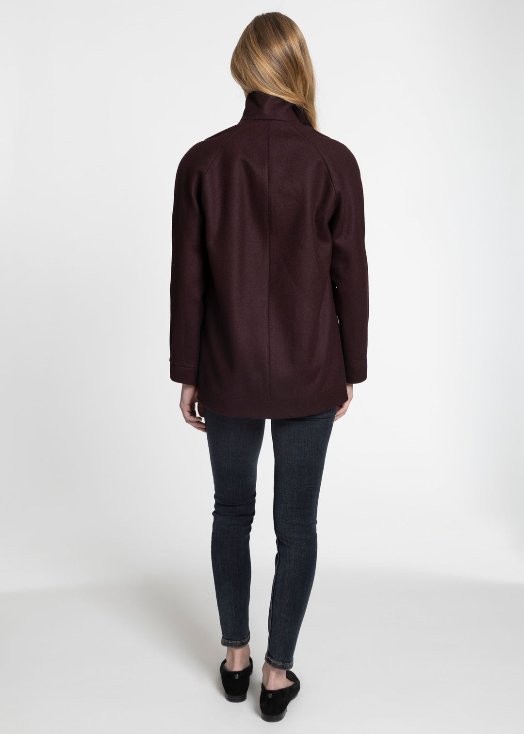 A woman viewed from the back wearing a dark burgundy long-sleeve jacket, dark gray skinny jeans, and black flats, standing against a plain white studio background.