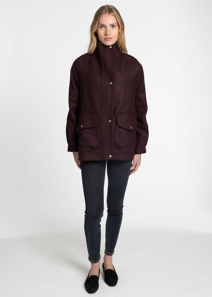 A woman modeling a dark burgundy utility jacket with a high collar, front button closure, and two front flap pockets, paired with dark skinny jeans and black slip-on shoes against a plain white studio background.