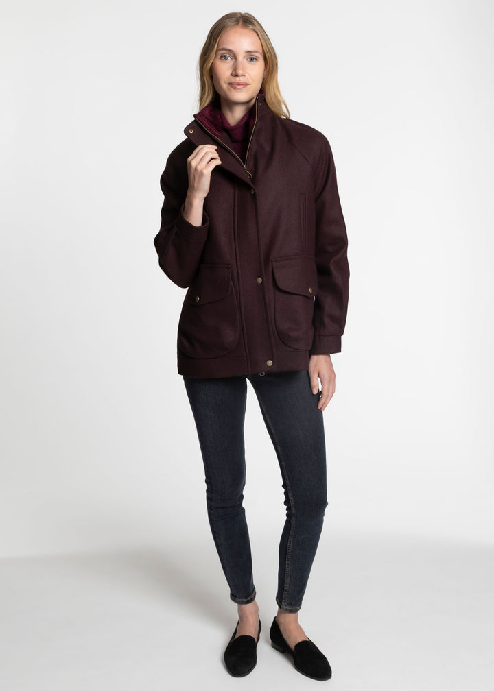 A woman modeling a dark plum-colored jacket with a high collar, front flap pockets, and gold-tone buttons, paired with dark gray skinny jeans and black slip-on shoes, against a plain white studio background.