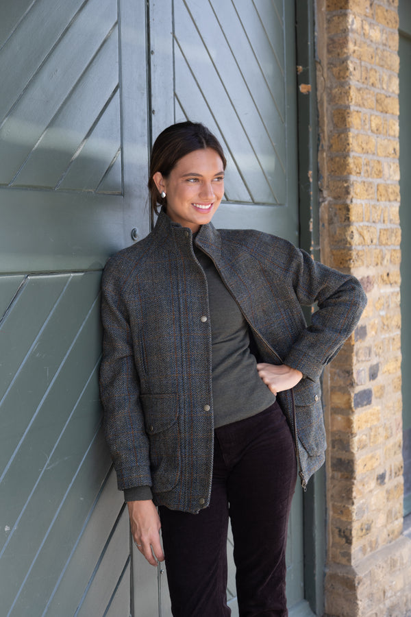 A woman wearing a green herringbone check wool coat with metal buttons, leaning against a door.