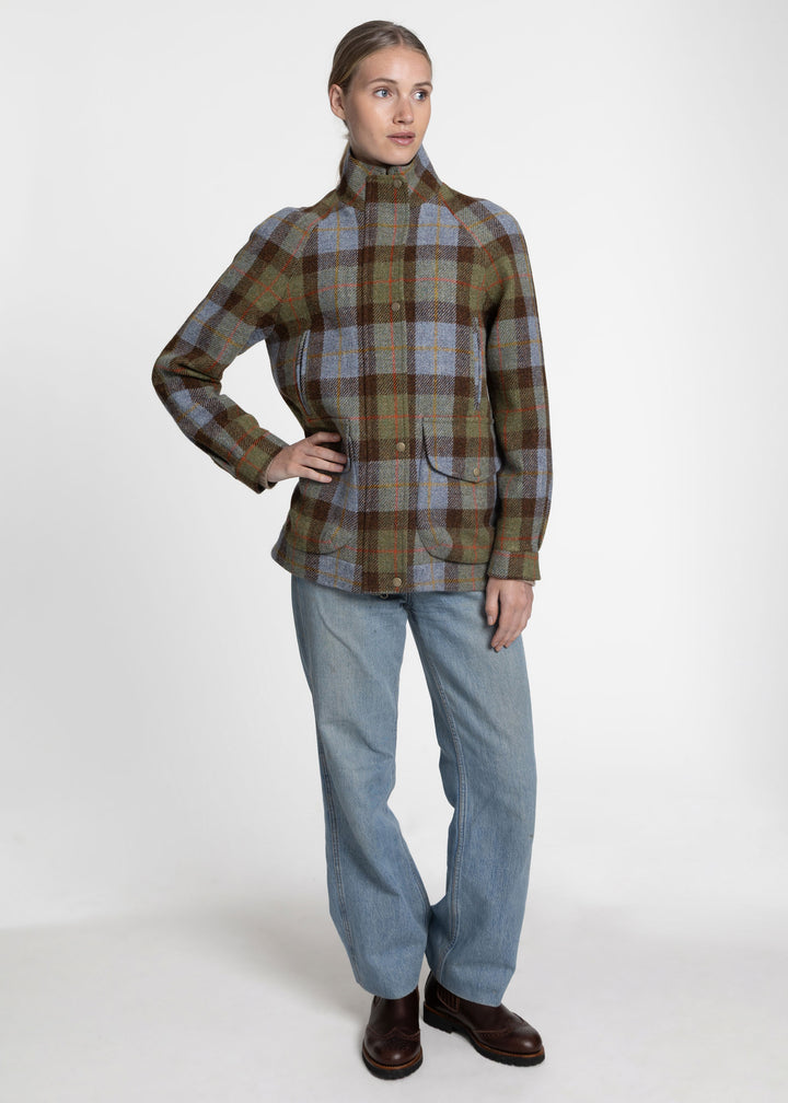 A woman modeling a high-neck, long-sleeve plaid jacket in green, brown, and blue tones, paired with light blue jeans and brown leather shoes, against a plain light gray studio background.