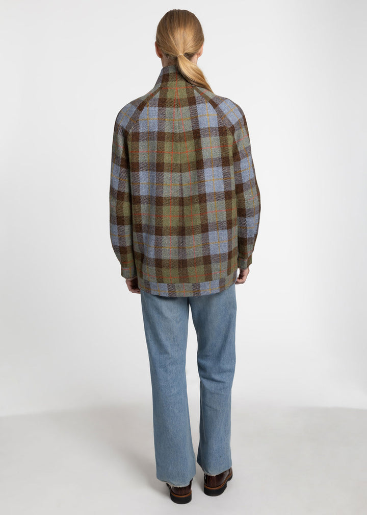 A woman viewed from behind wearing a brown, blue, and green plaid jacket with light blue jeans and brown shoes, standing against a plain white studio background.