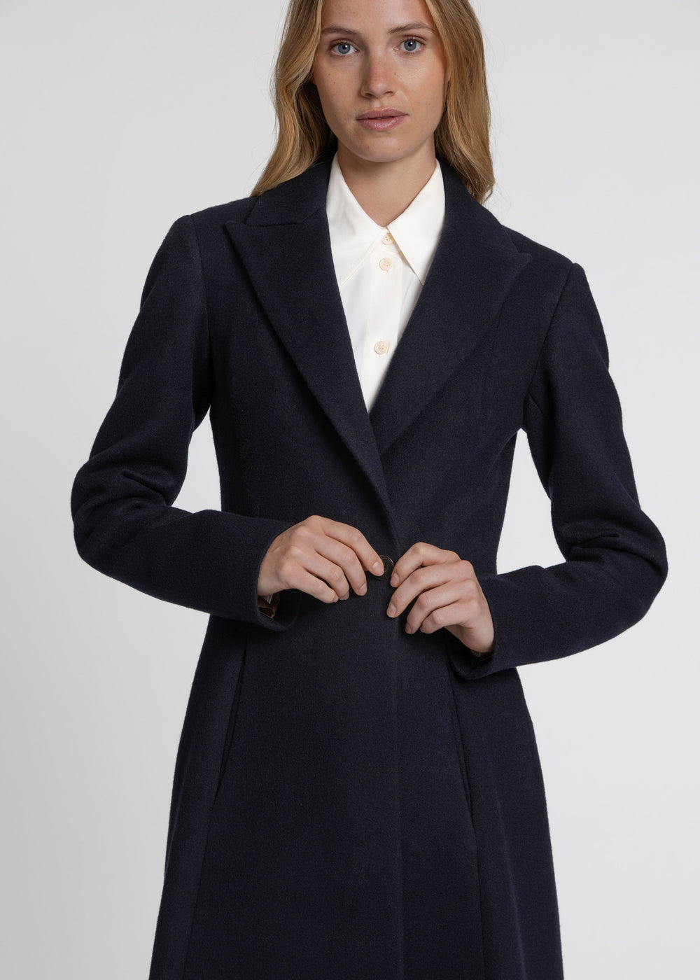 A woman wearing a dark navy wool-blend coat over a white collared shirt, photographed against a light gray studio background.