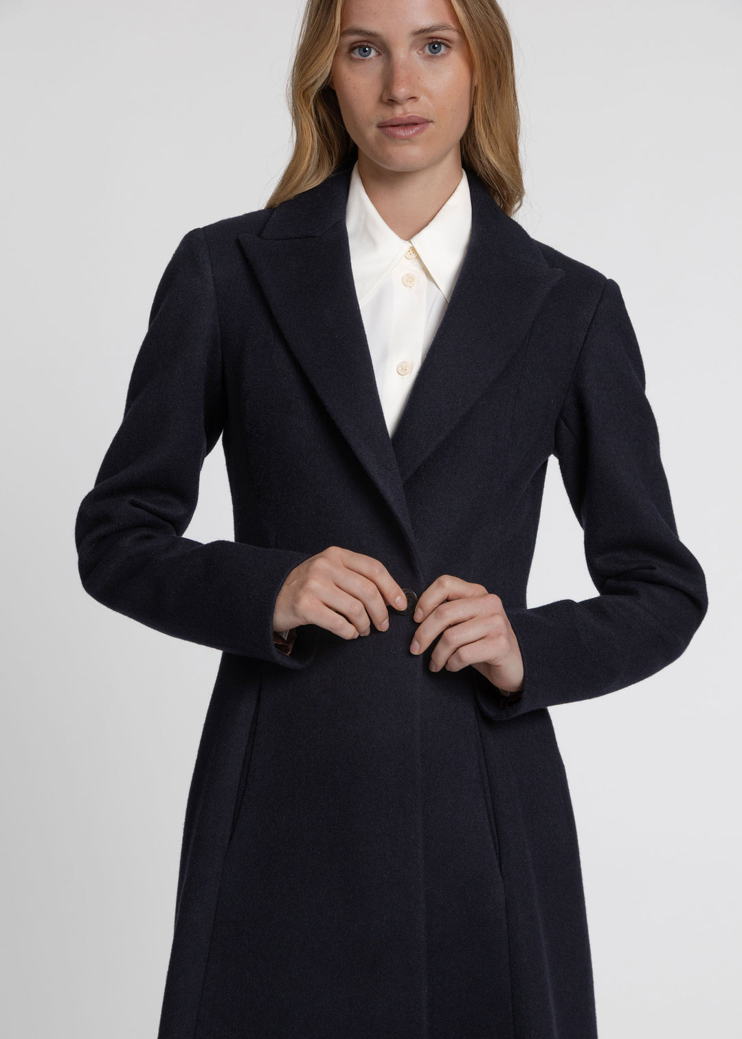 A woman wearing a dark navy wool-blend coat over a white collared shirt, photographed against a light gray studio background.