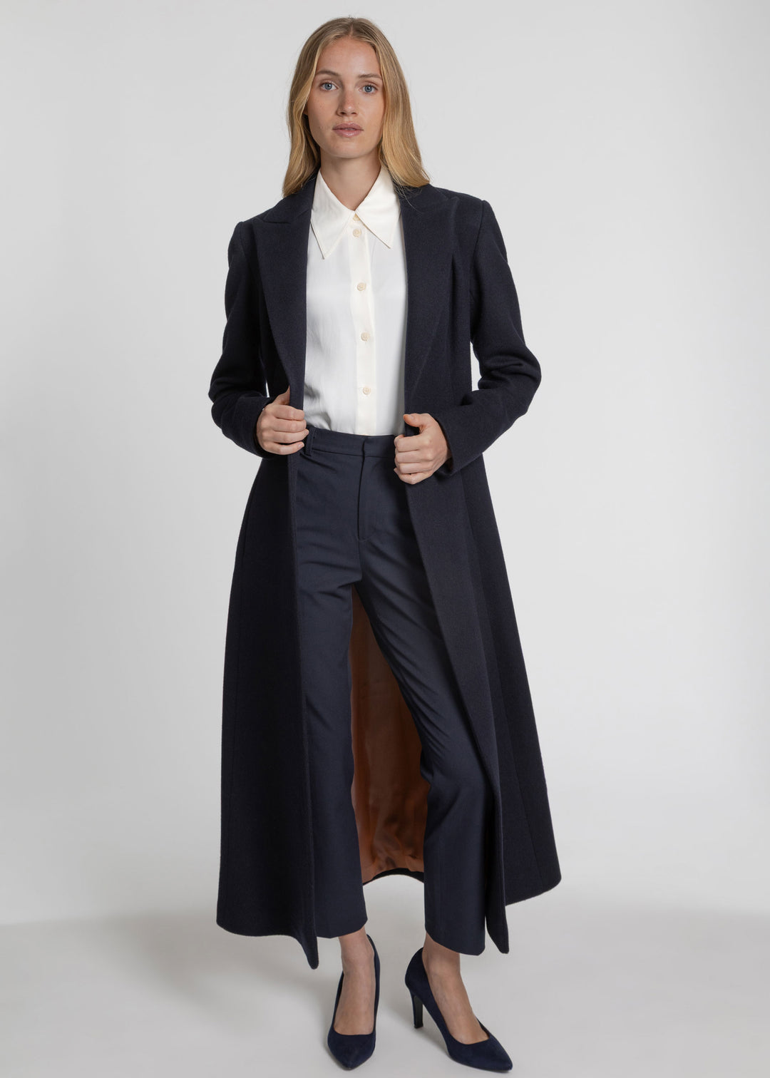 A woman models a long navy blue tailored overcoat over a white collared blouse, matching navy trousers, and navy heels against a plain light gray studio background.