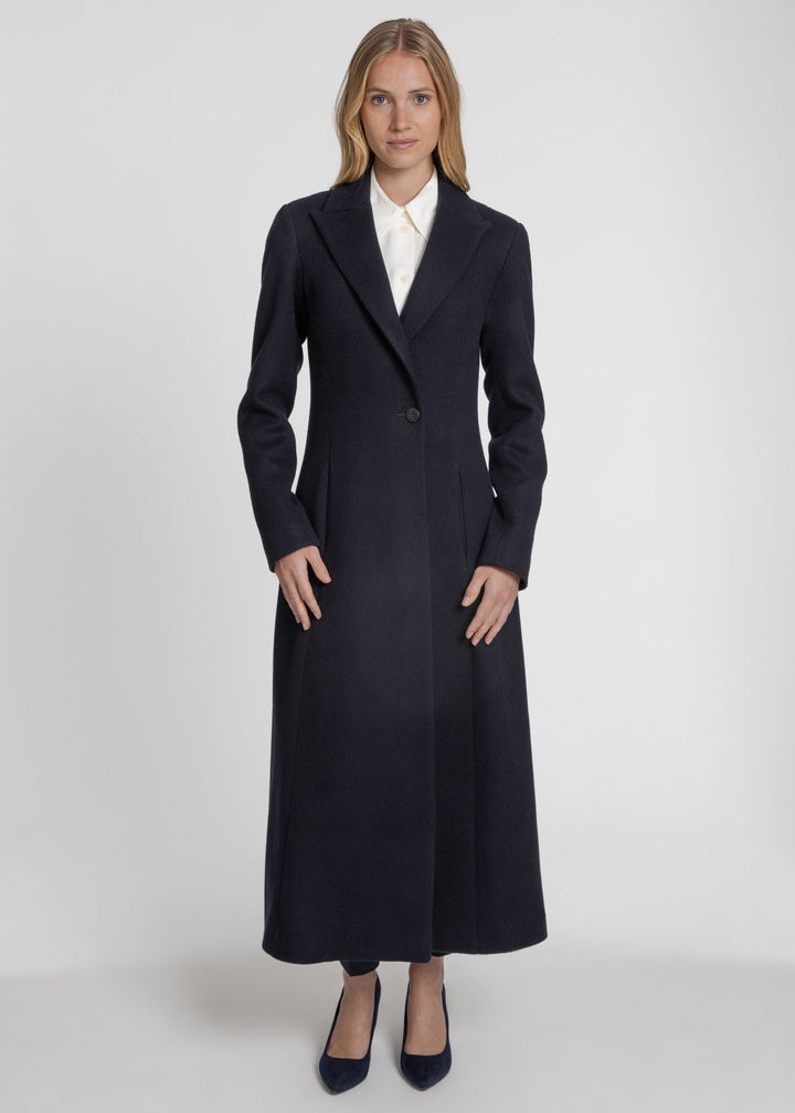 A woman modeling a long, dark navy wool-blend coat with a single front button, worn over a white collared shirt and navy heels, against a light gray studio background.