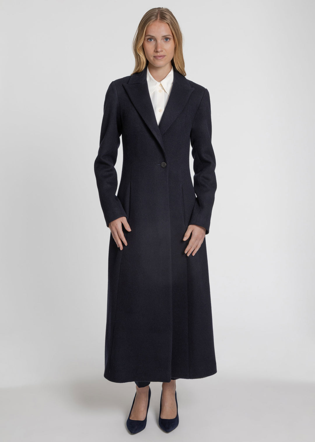 A woman modeling a long, dark navy wool-blend coat with a single front button, worn over a white collared shirt and navy heels, against a light gray studio background.
