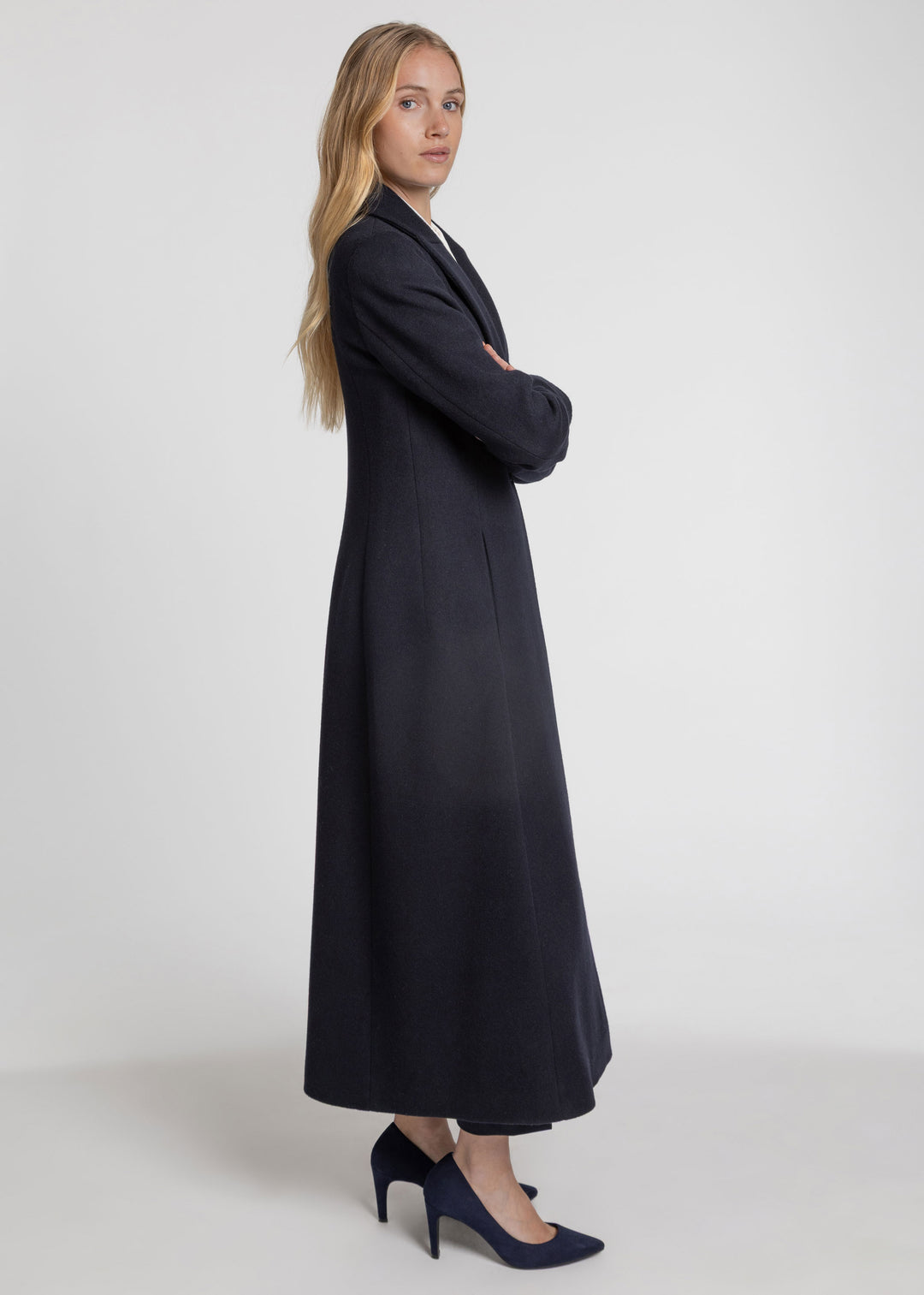 A woman modeling a long, dark navy blue coat with a structured collar and long sleeves, paired with navy blue heels, against a plain light gray studio background.