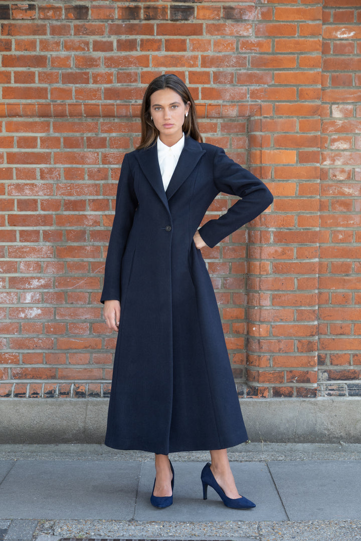 A woman stands against a red brick wall wearing a long navy blue coat over a white top, paired with navy blue heels.