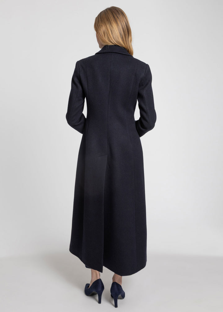 A woman viewed from the back wearing a long dark navy coat and navy heels against a light gray studio background.