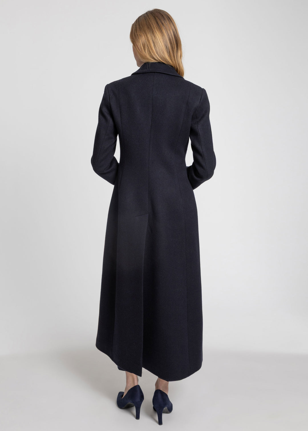 A woman viewed from the back wearing a long dark navy coat and navy heels against a light gray studio background.
