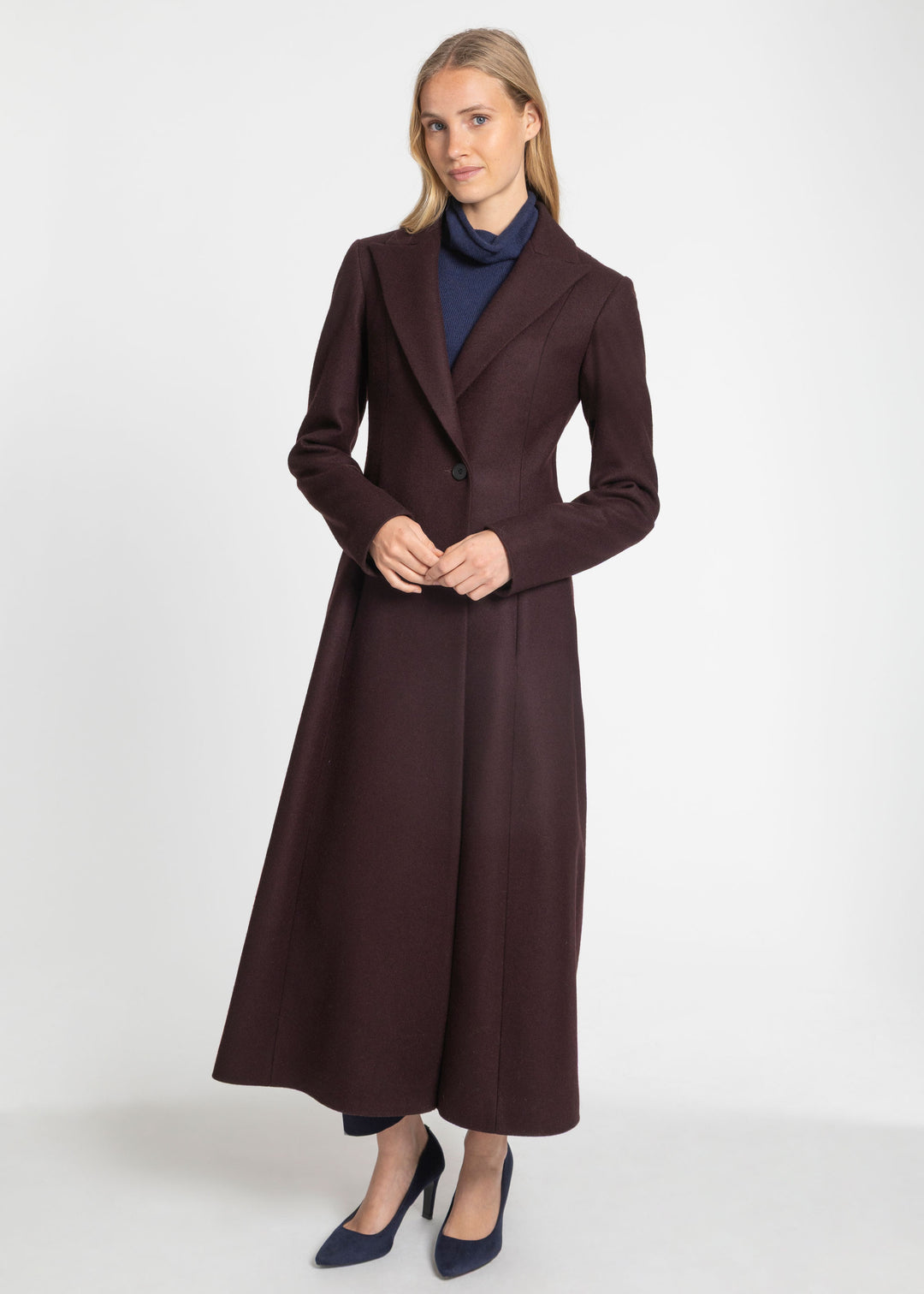 A woman modeling a long, dark mulberry tailored coat over a navy blue turtleneck sweater and navy blue heels, standing against a plain light gray studio background.