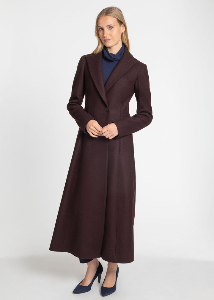 A woman modeling a long, dark mulberry tailored coat over a navy blue turtleneck sweater and navy blue heels, standing against a plain light gray studio background.