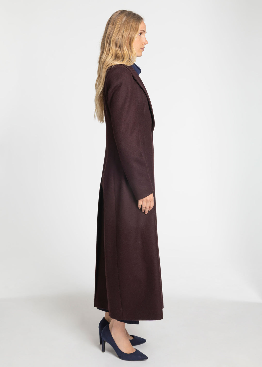 A woman in profile wearing a long, dark mulberry wool-blend coat over a navy blue top and navy blue heels, standing against a plain white studio background.