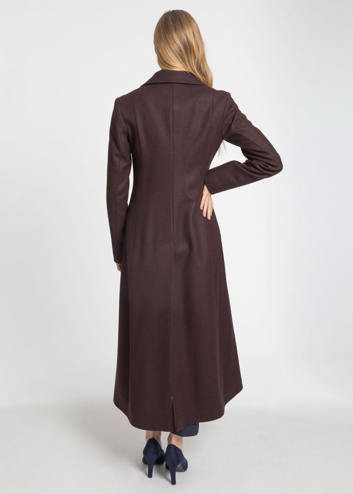 A woman viewed from the back wearing a long dark mulberry wool coat, paired with dark blue heels, against a plain light gray studio background.