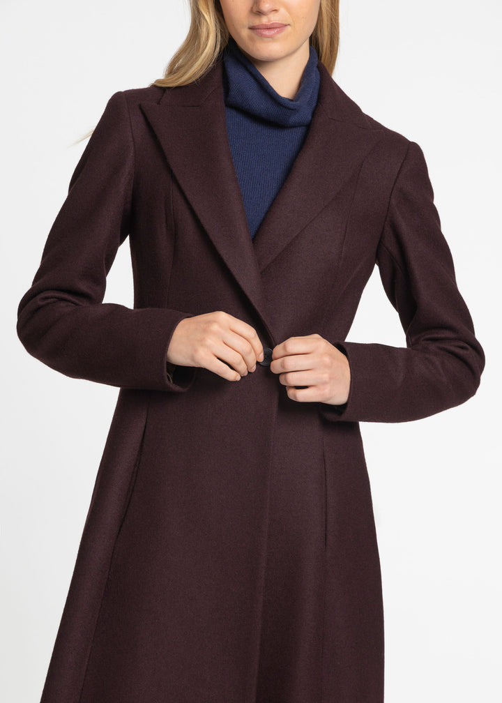 A woman wearing a dark plum-brown wool coat over a navy blue turtleneck sweater, shown from the chin down against a plain light gray studio background.