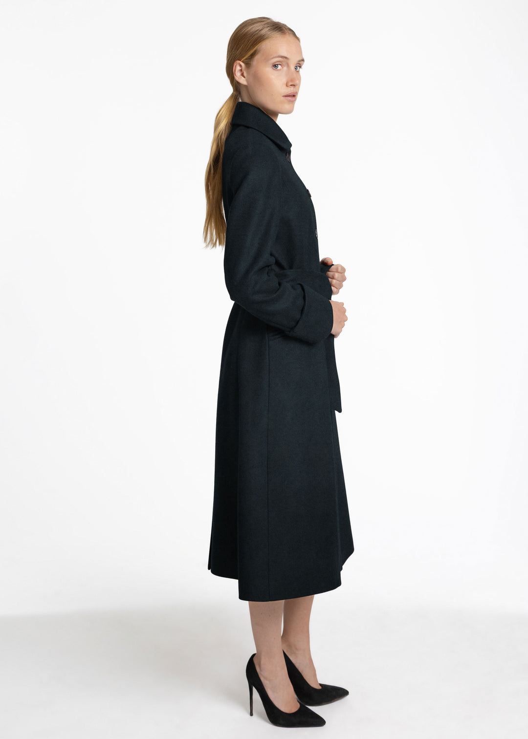 A woman modeling a long green coat with a collar and long sleeves, paired with black high heels, photographed against a plain white studio background.