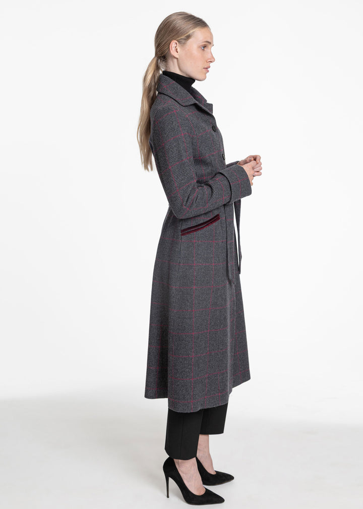 A woman modeling a long gray windowpane plaid coat with a black turtleneck and black trousers, shown from a side profile against a white studio background.