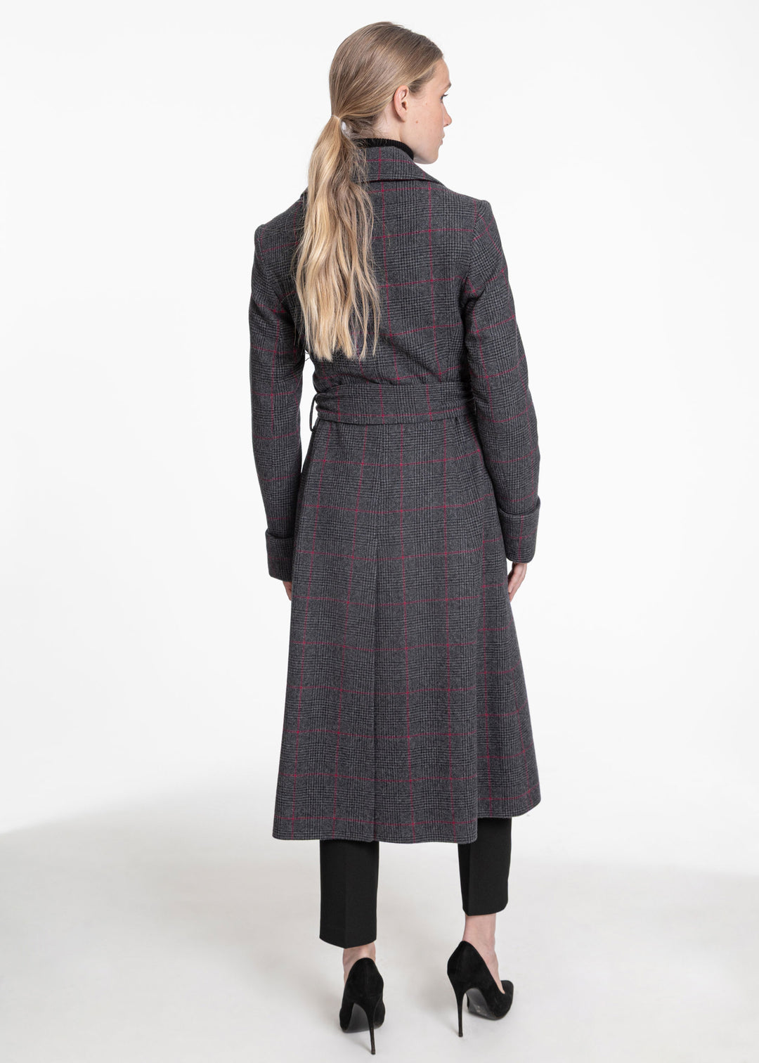 A woman viewed from the back wearing a long dark gray windowpane coat with thin red lines, paired with black pants and black heels, against a plain white studio background.