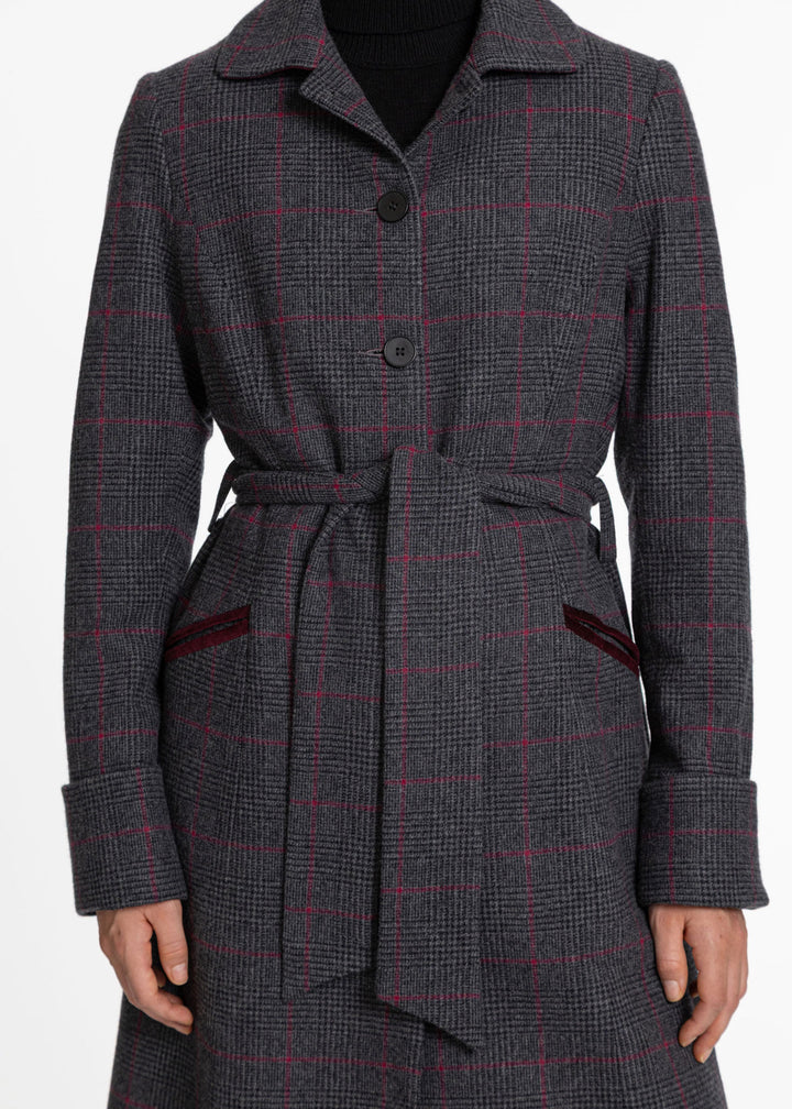 A close up of a woman wearing a black and grey check coat with a pinkish red overcheck. Buttons and belted closure with burgundy velvet pocket trim.