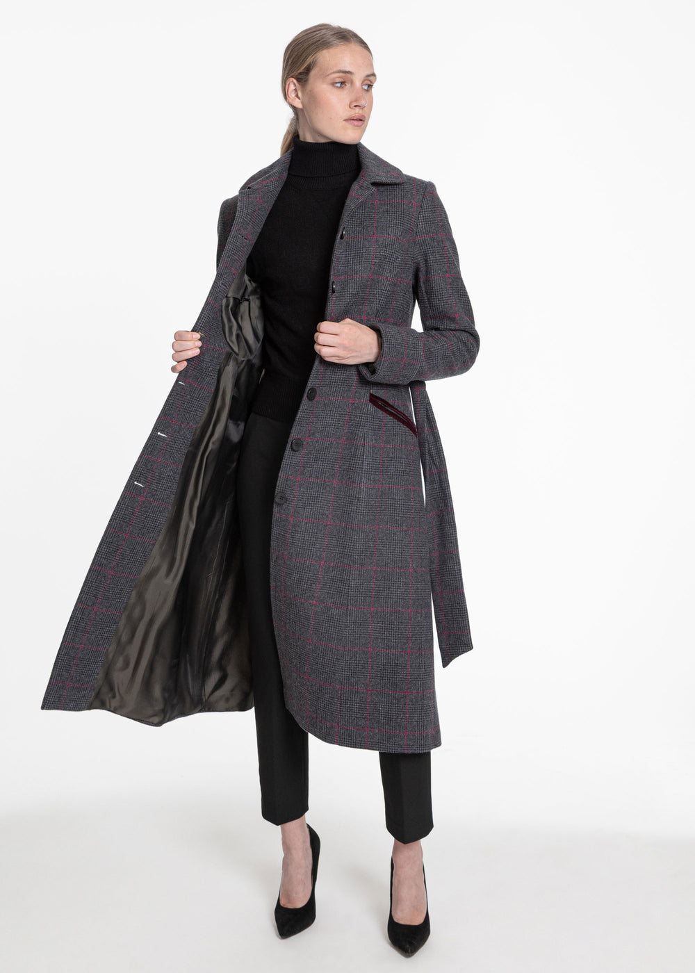 A woman models a long gray windowpane plaid coat with red lines, worn open over a black turtleneck and black pants, against a plain white studio background.