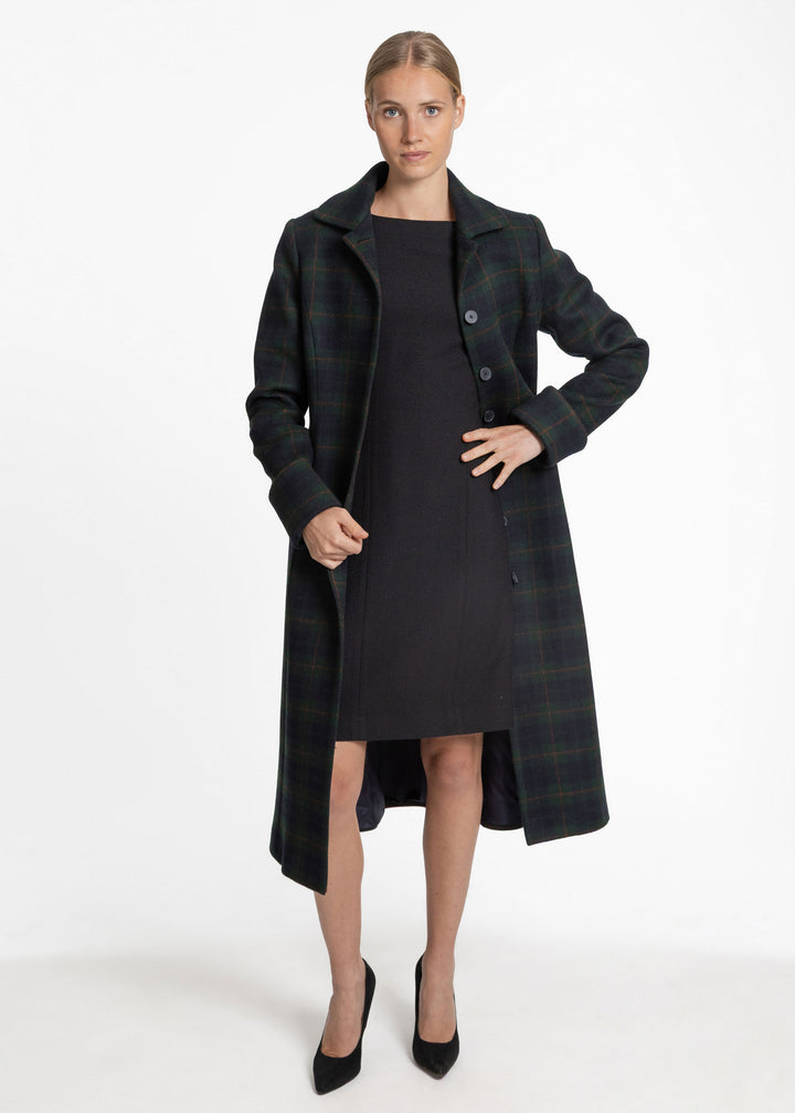 A woman modeling a long dark plaid coat over a black dress and black pointed heels, photographed against a clean white studio background.