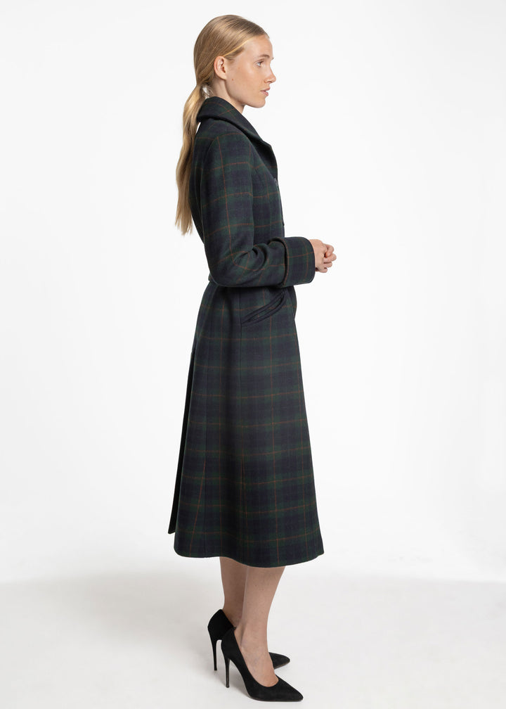 A woman in profile wearing a long dark green and navy plaid coat with a collar, paired with black high heels, photographed against a plain white studio background.