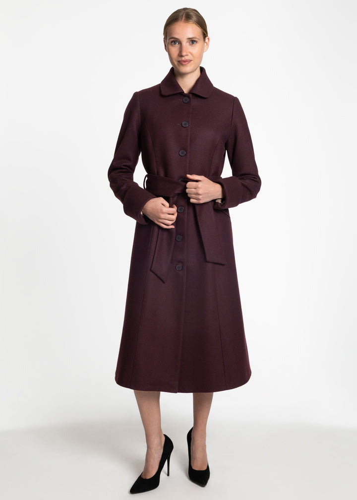 A woman modeling a long, dark plum-burgundy coat with a collar, front buttons, and a matching waist tie, paired with black high heels against a plain light gray studio background.