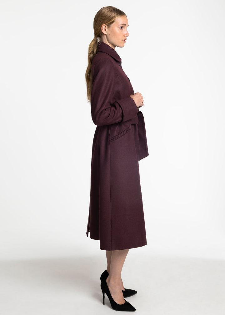 A woman in profile wearing a long, dark plum-burgundy coat, styled with black heels against a plain white studio background.